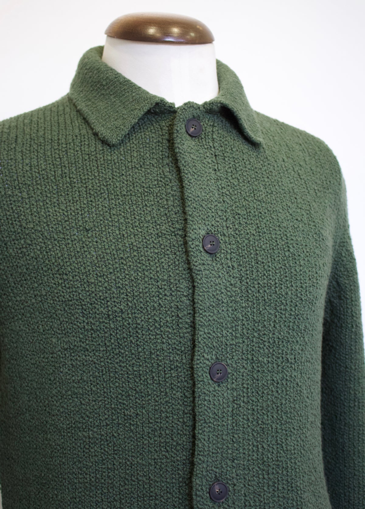 A dark green textured knit cardigan sweater with a collar and dark buttons, displayed on a white mannequin torso with a brown wooden neck cap.