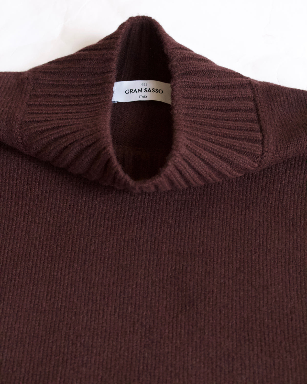 Close-up of a ribbed knit turtleneck sweater in a deep burgundy brown, showing the collar and a white interior label that reads 'GRAN SASSO'.