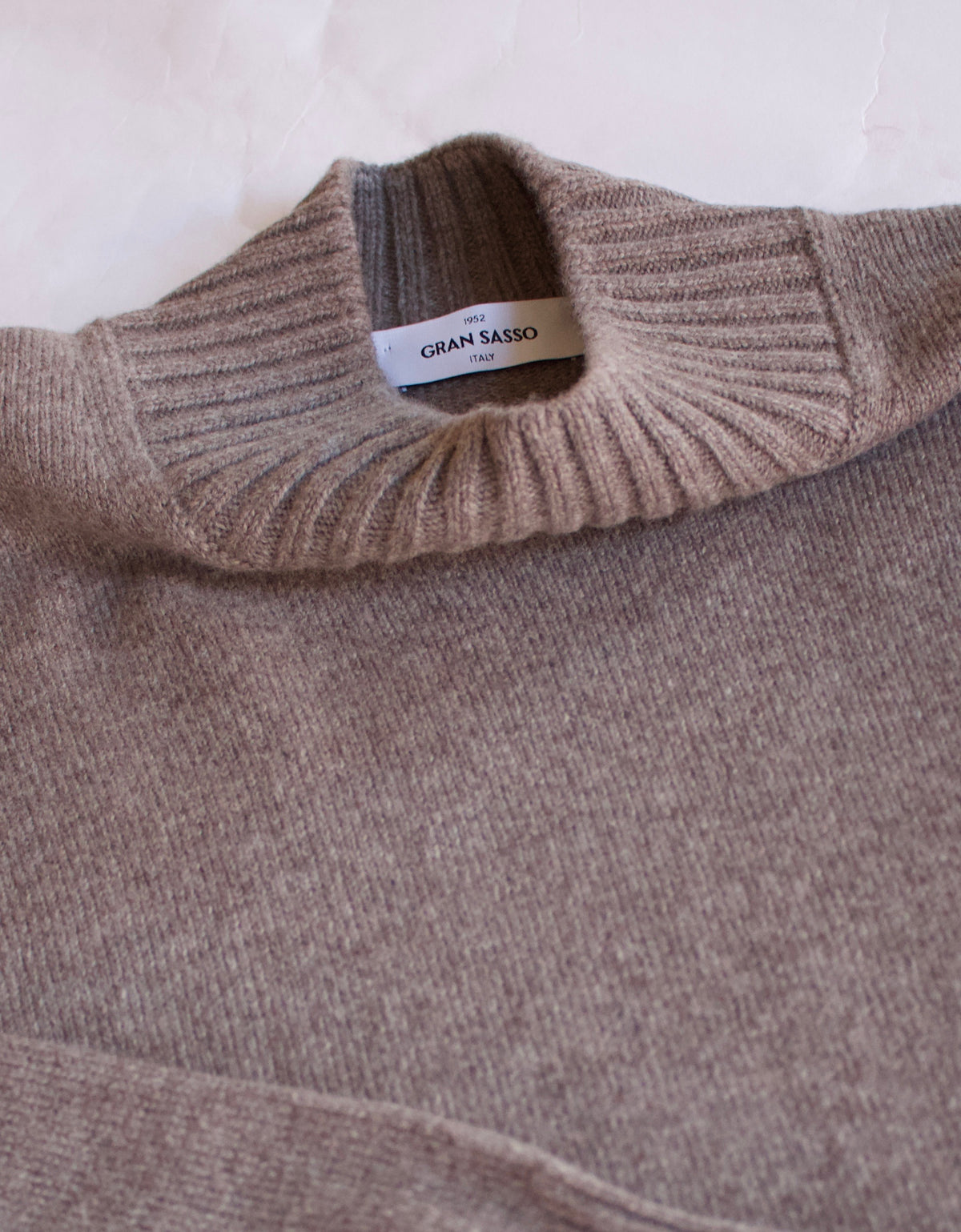Close-up of a ribbed knit turtleneck sweater in a warm taupe-brown color, showing the interior neck label which reads GRAN SASSO.