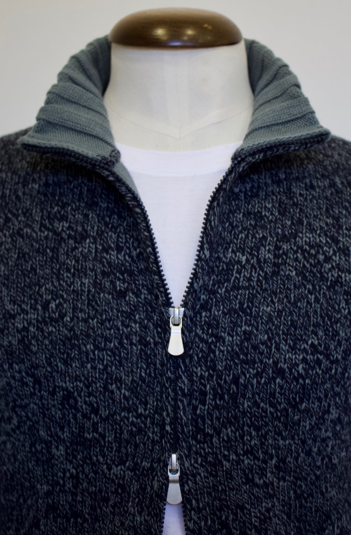 A close-up of a dark heathered zip-up sweater displayed on a mannequin torso, layered over a white shirt. The vest features a ribbed collar in a muted pale blue tone and a silver zipper.