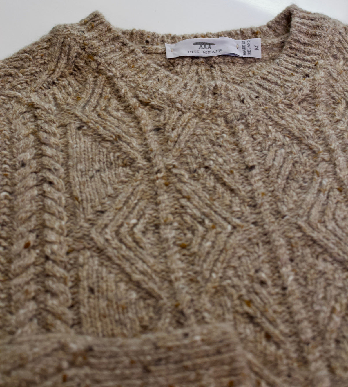Photo of a creamy Aran sweater flat laid on a white background. You can see the label of the knitwear brand Inis Meain and a Made In Ireland label on the inside collar. The sweater has a multi toned cable pattern in beiges, creams and browns.
