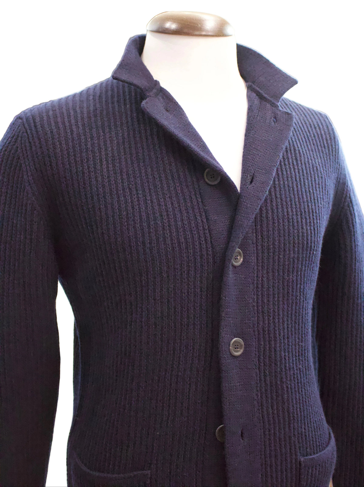 A photo of a navy ribbed heavy cardigan from the Irish knitwear brand Inis Meain on a white a background. The cardigan has a nehru collar and a wide thick placket, horn buttons up the front and two front pockets.