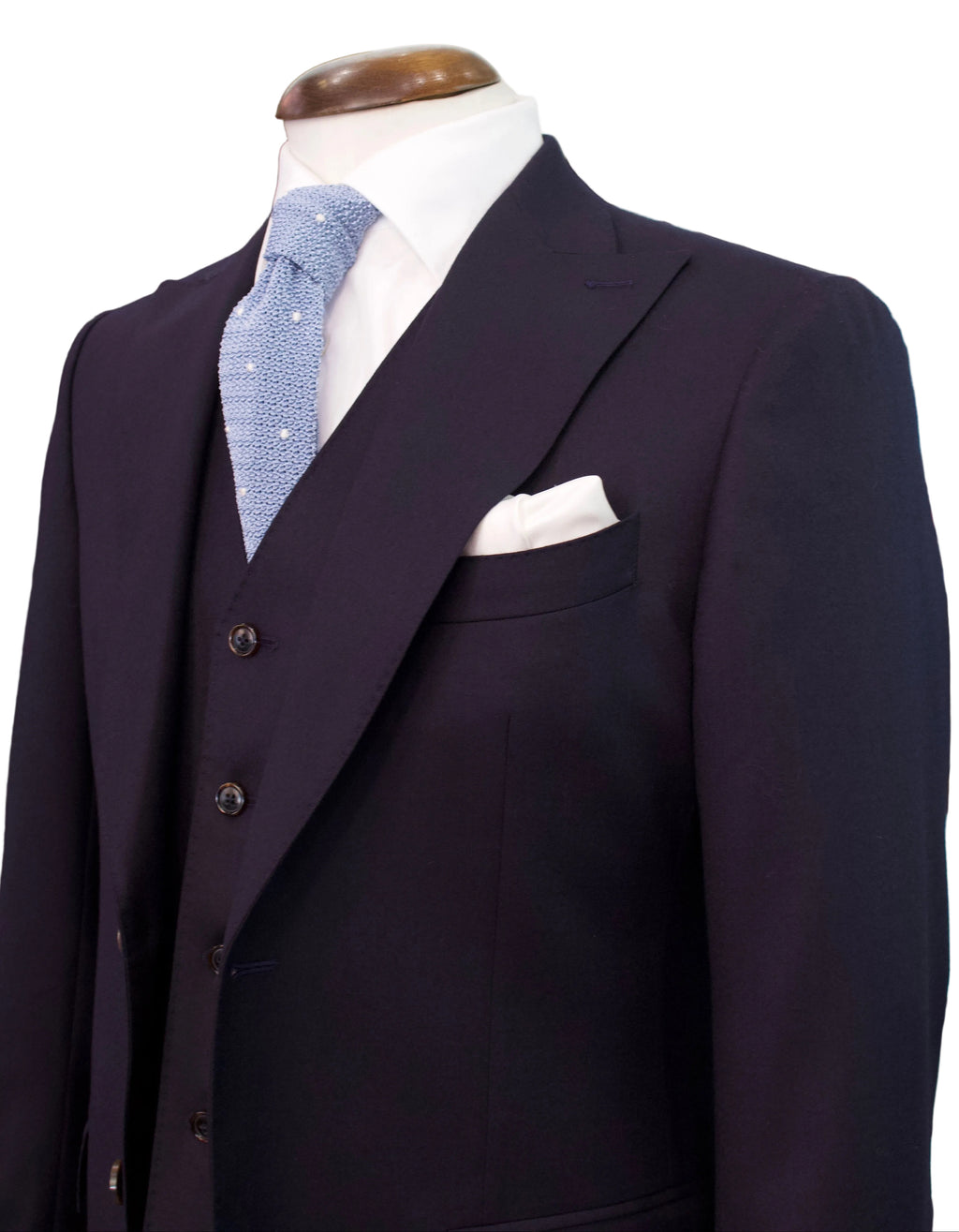 A dark navy three-piece suit displayed on a mannequin torso, styled with a light blue patterned tie, a white pocket square, and a white dress shirt in a retail showroom.