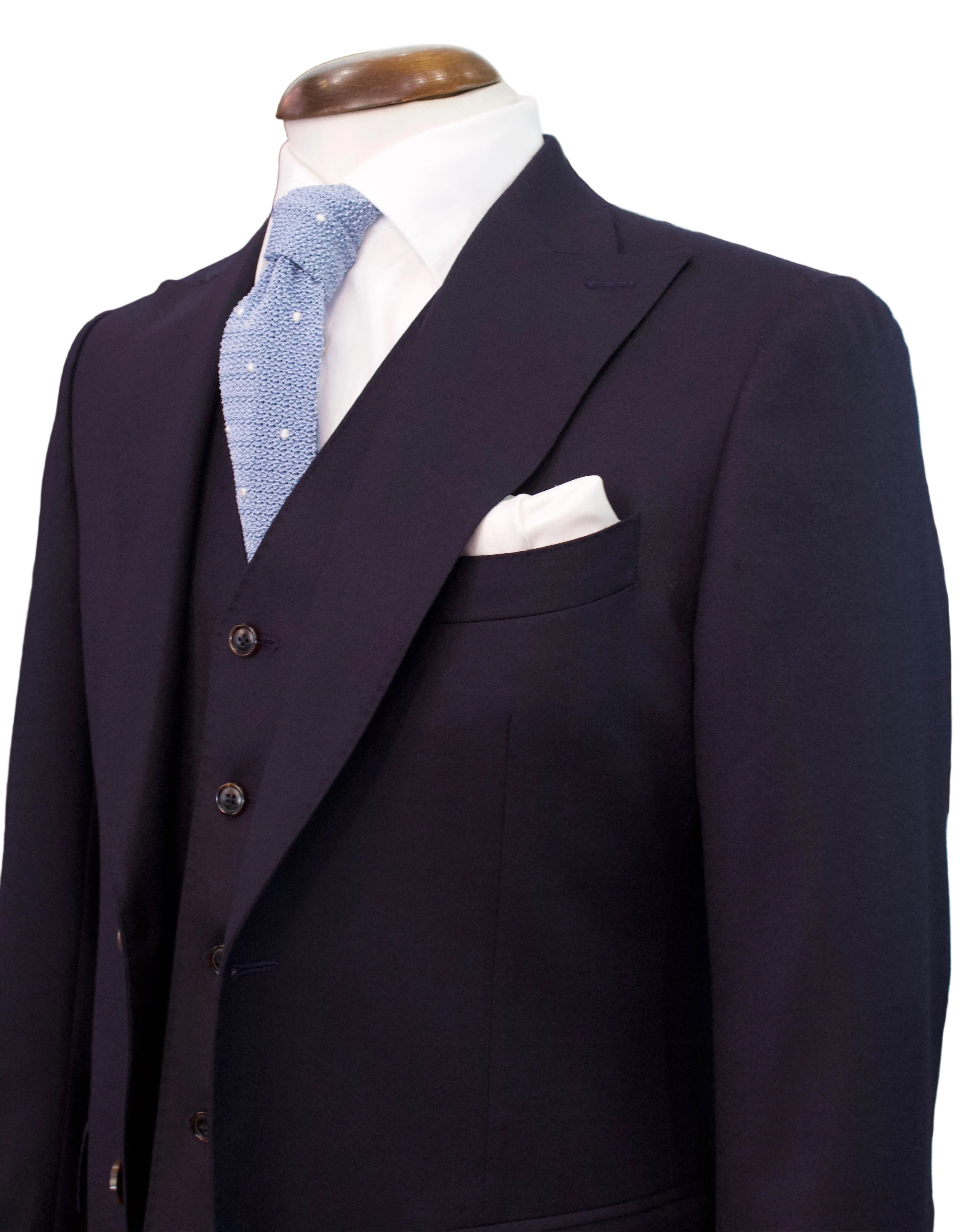 A dark navy three-piece suit displayed on a mannequin torso, styled with a light blue patterned tie, a white pocket square, and a white dress shirt in a retail showroom.