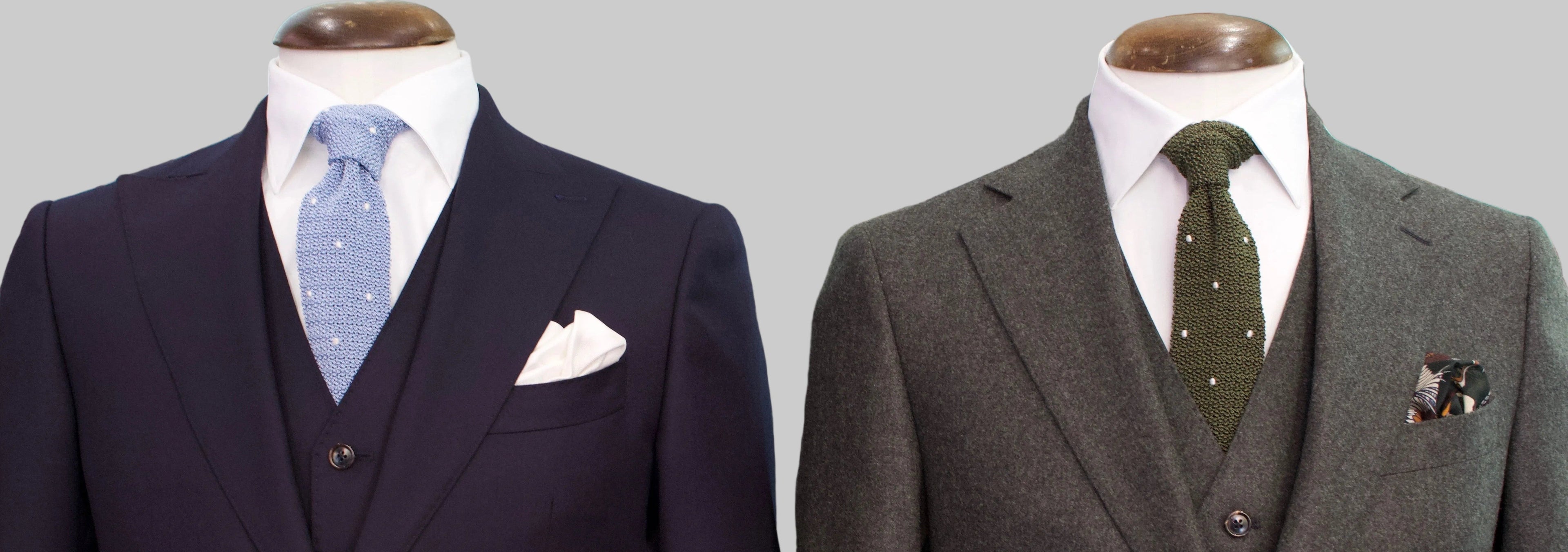 Two men's three-piece suits displayed on mannequins in a showroom. The left suit is dark navy blue with a light blue patterned tie and white pocket square.