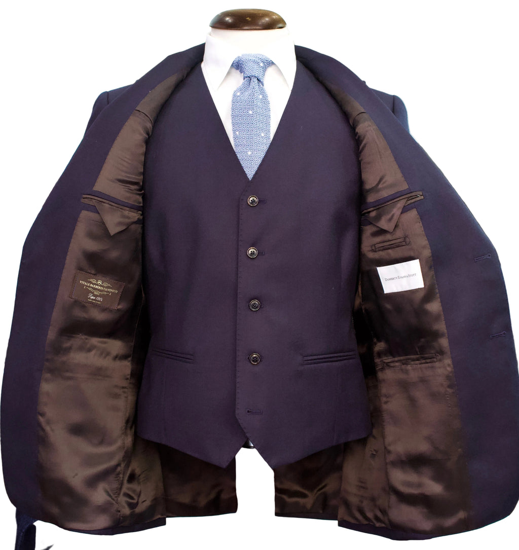 A men's three-piece suit displayed on a mannequin torso, featuring a deep purple vest, matching trousers, and a light blue patterned tie. The suit jacket is open to show a dark brown satin-lined interior, set against a softly blurred boutique background.